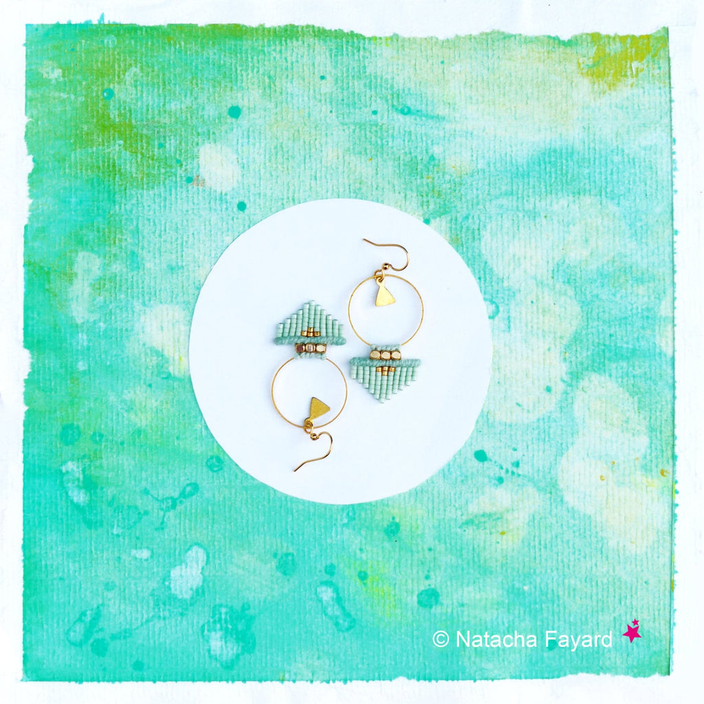 Peut inclure: Une paire de boucles d'oreilles en perles bleu clair et or avec un motif géométrique. Les boucles d'oreilles sont suspendues à une surface circulaire blanche sur un fond aquarelle vert.