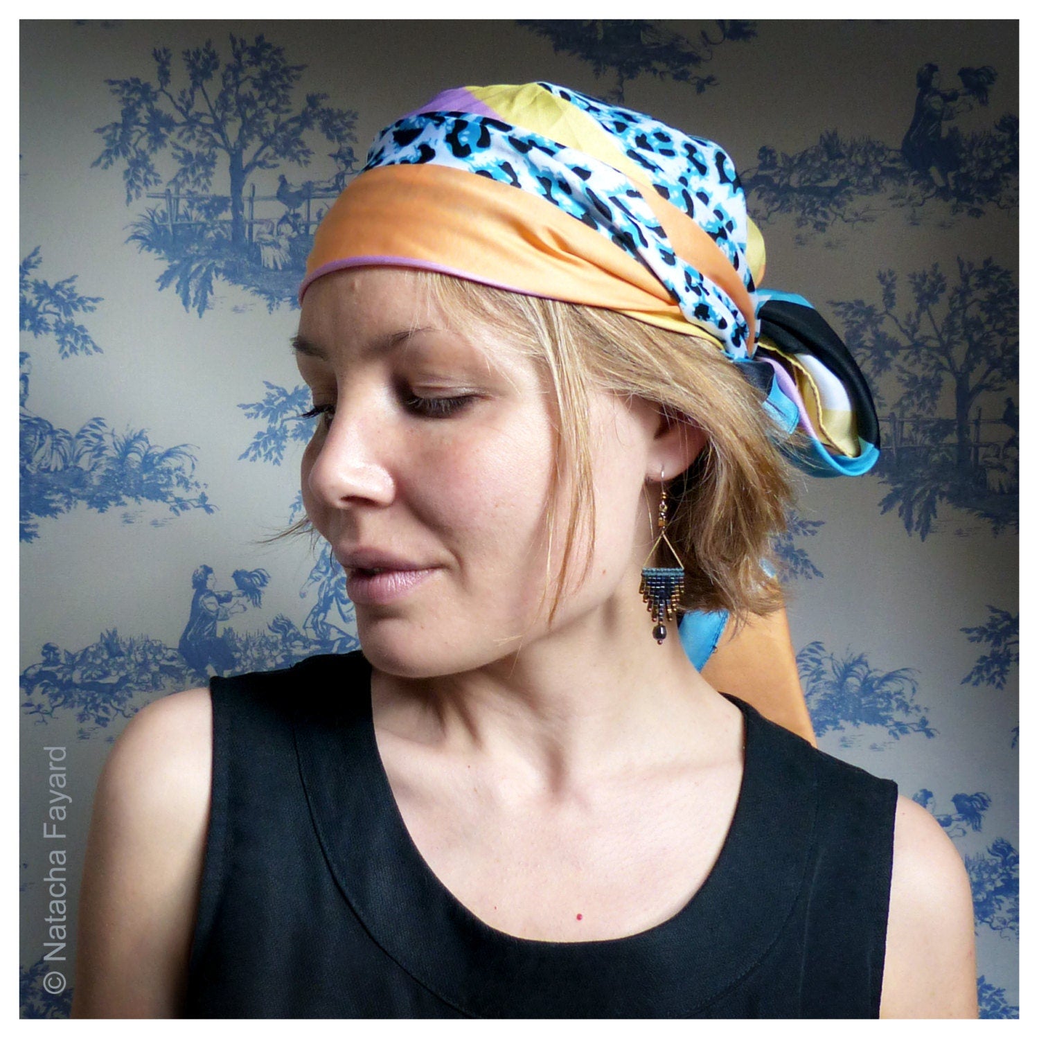 Peut inclure: Une femme aux cheveux blonds porte un foulard à motifs colorés et un haut noir sans manches. Elle porte une paire de boucles d'oreilles pendantes avec un motif géométrique.