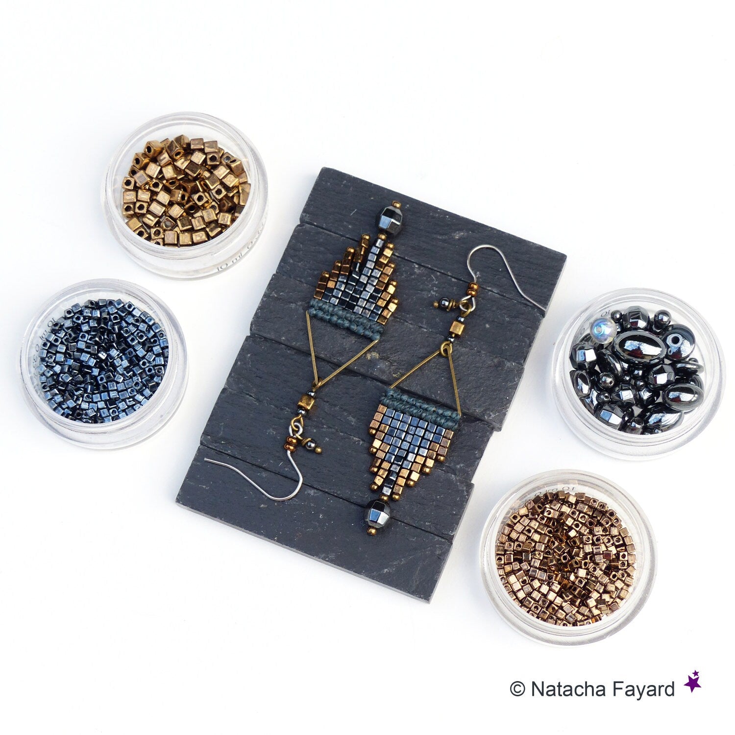 Peut inclure: Une paire de boucles d'oreilles faites à la main avec un design géométrique. Les boucles d'oreilles sont faites avec des perles dorées, noires et bleues. Les boucles d'oreilles sont présentées sur une surface en ardoise noire avec de petits récipients de perles.