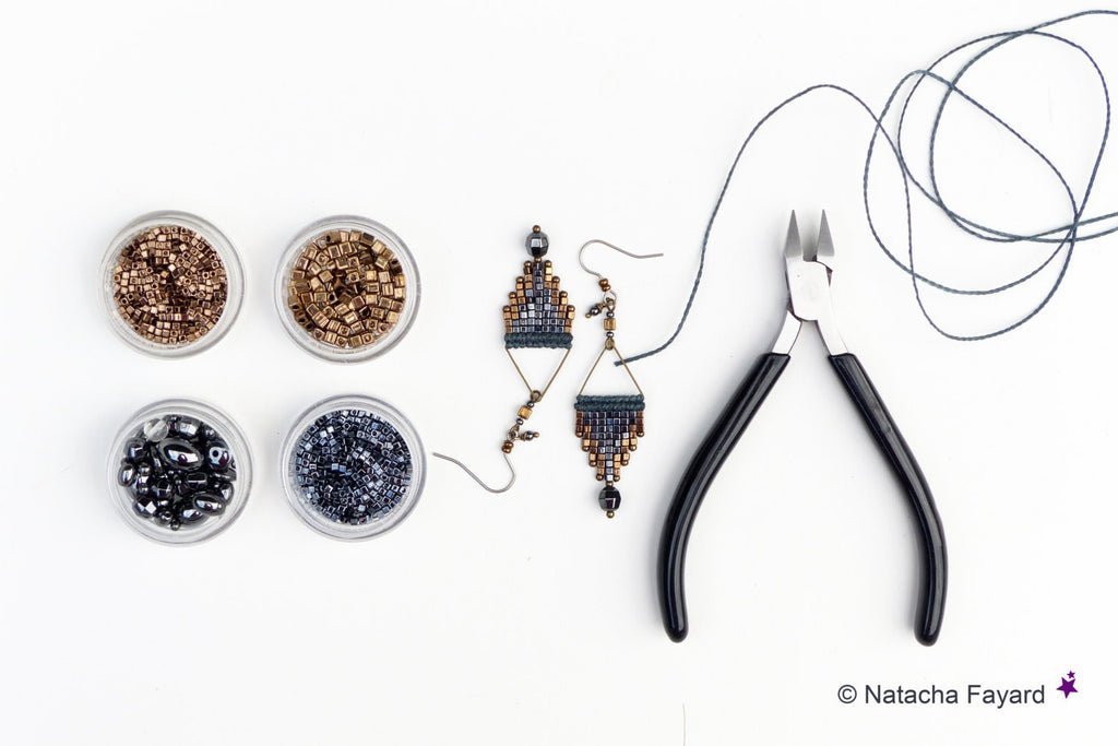 Peut inclure: Deux paires de boucles d'oreilles en perles avec un motif géométrique dans des tons de bleu, noir et or. Les boucles d'oreilles sont présentées sur une surface blanche avec de petits contenants de perles, une bobine de fil et des pinces coupantes.