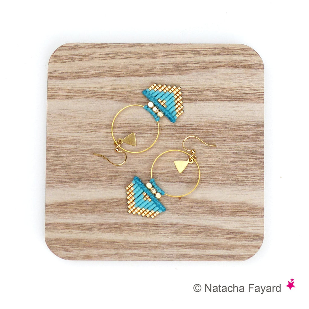 Peut inclure: Une paire de boucles d'oreilles créoles dorées avec des franges de perles turquoise et dorées qui pendent du bas. Les boucles d'oreilles sont sur une surface en bois.