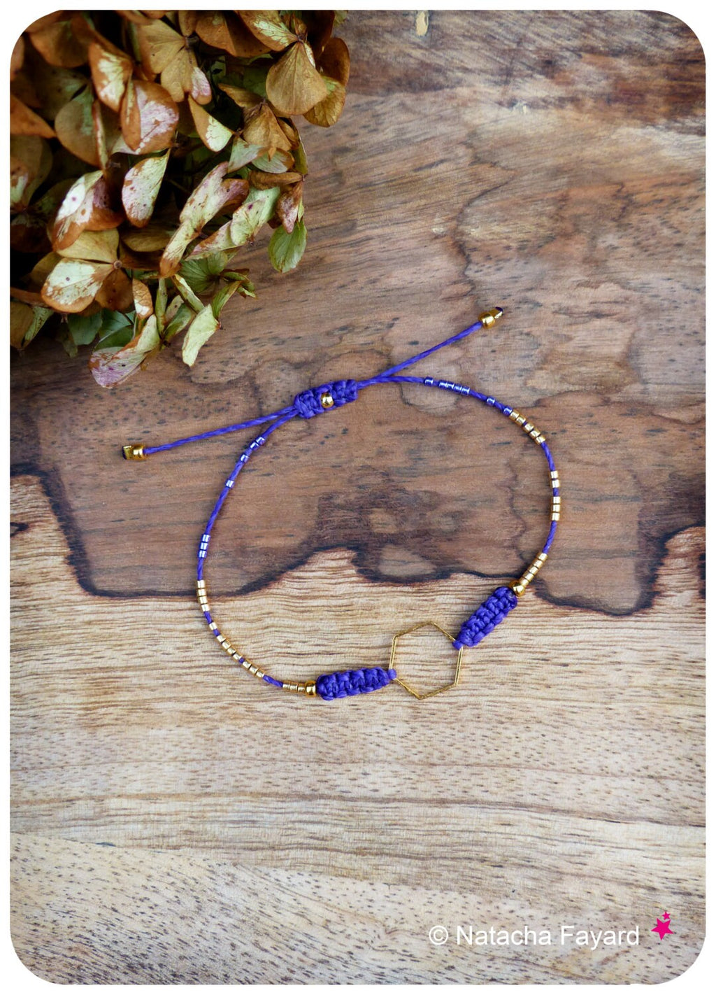 Peut inclure: Bracelet fait main avec un charm hexagonal doré et des perles dorées. Le bracelet est composé d'un cordon bleu avec des nœuds coulissants réglables. Le design est posé sur un fond en bois, avec des feuilles séchées en haut à gauche.