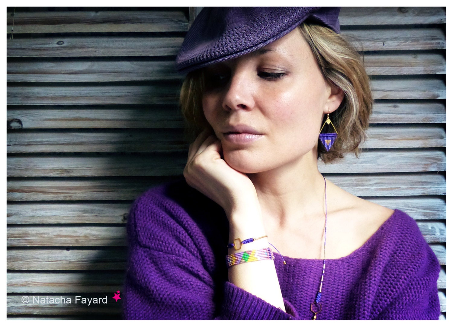 Peut inclure: Une femme porte un béret et un pull violets, avec des bijoux de perles. Les boucles d'oreilles sont triangulaires avec des accents dorés et des perles bleues. Un bracelet avec des perles colorées est également visible. L'arrière-plan est un volet en bois.