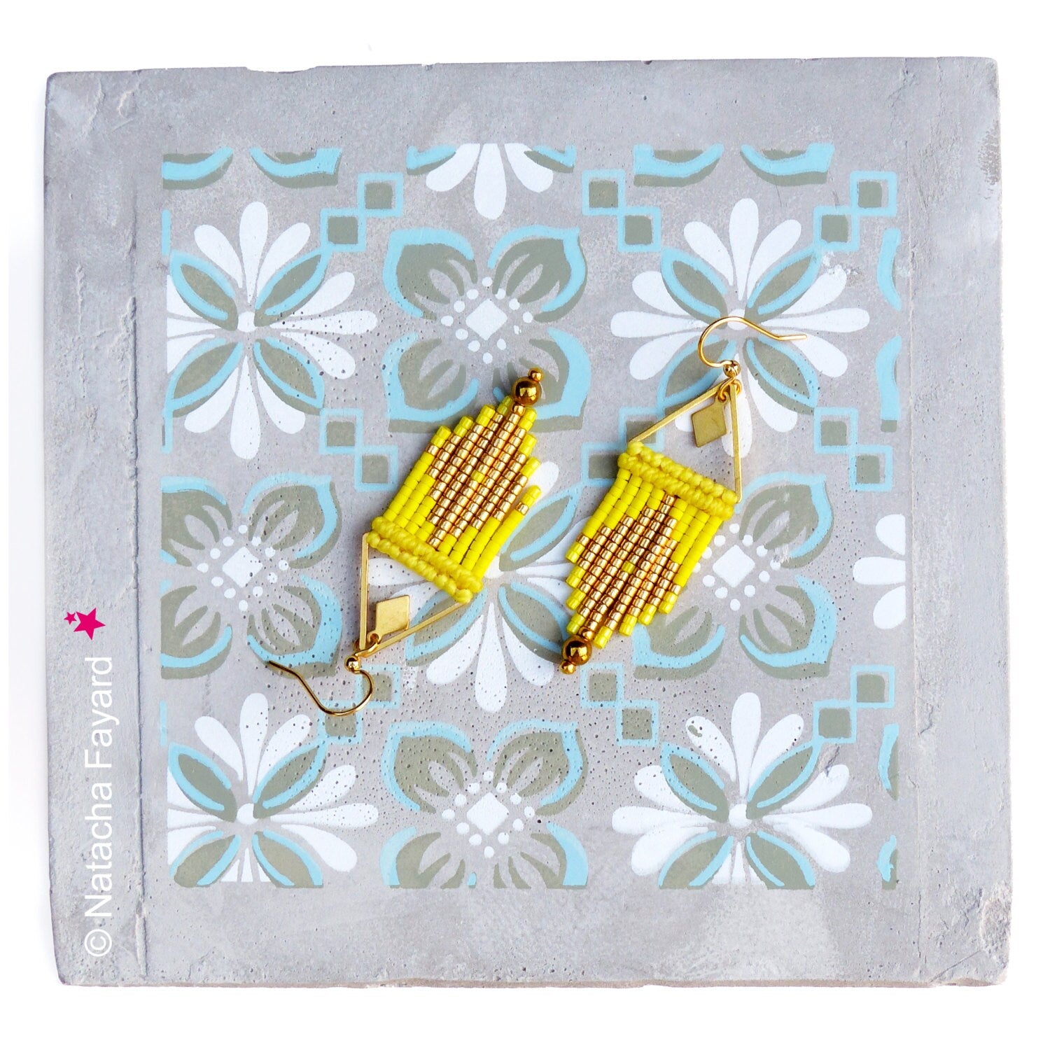 Peut inclure: Une paire de boucles d'oreilles faites à la main, jaunes et dorées, avec un motif géométrique. Les boucles d'oreilles ont une forme triangulaire et des accents en métal doré. Elles sont présentées sur une surface grise à motifs.