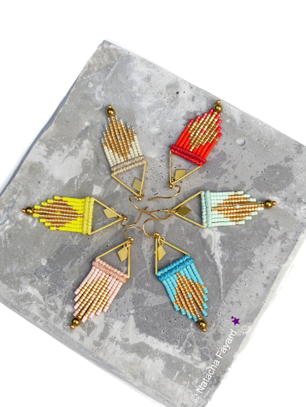Peut inclure: Six paires de boucles d'oreilles en perles colorées avec des accents de triangle dorés. Les boucles d'oreilles sont disposées en forme d'étoile sur une surface en béton gris.