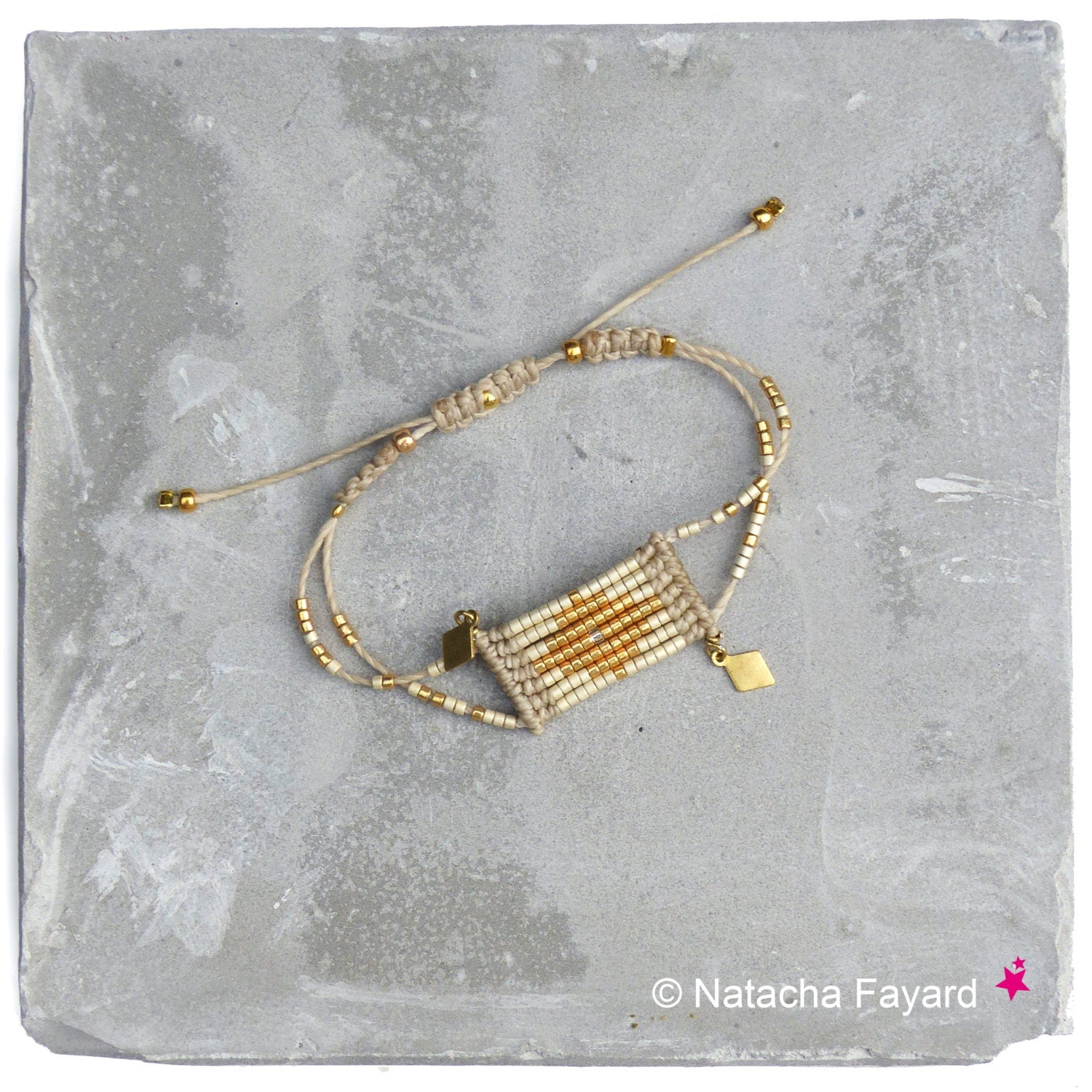 Peut inclure: Un bracelet en perles beige et or avec un motif tissé et deux breloques dorées. Le bracelet est sur un fond gris.