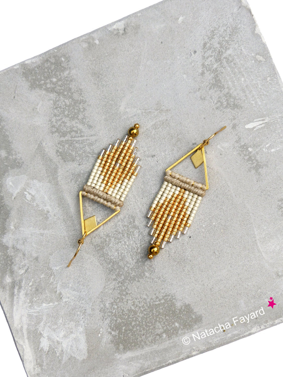 Peut inclure: Une paire de boucles d'oreilles en perles dorées et blanches avec un motif géométrique. Les boucles d'oreilles sont suspendues à un fil doré et ont une petite perle dorée au bas.