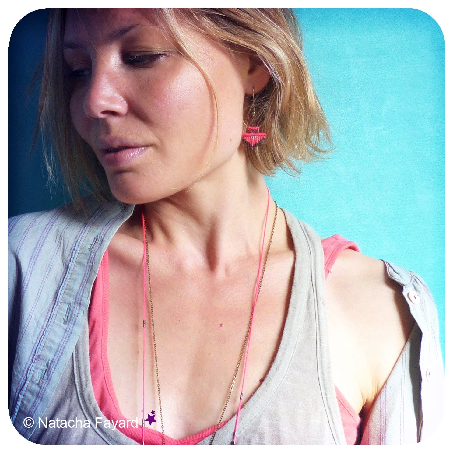 Peut inclure: Une femme portant une chemise gris clair avec un débardeur rose en dessous. Elle porte un collier de chaîne en or et des boucles d'oreilles roses. L'arrière-plan est un mur bleu clair.