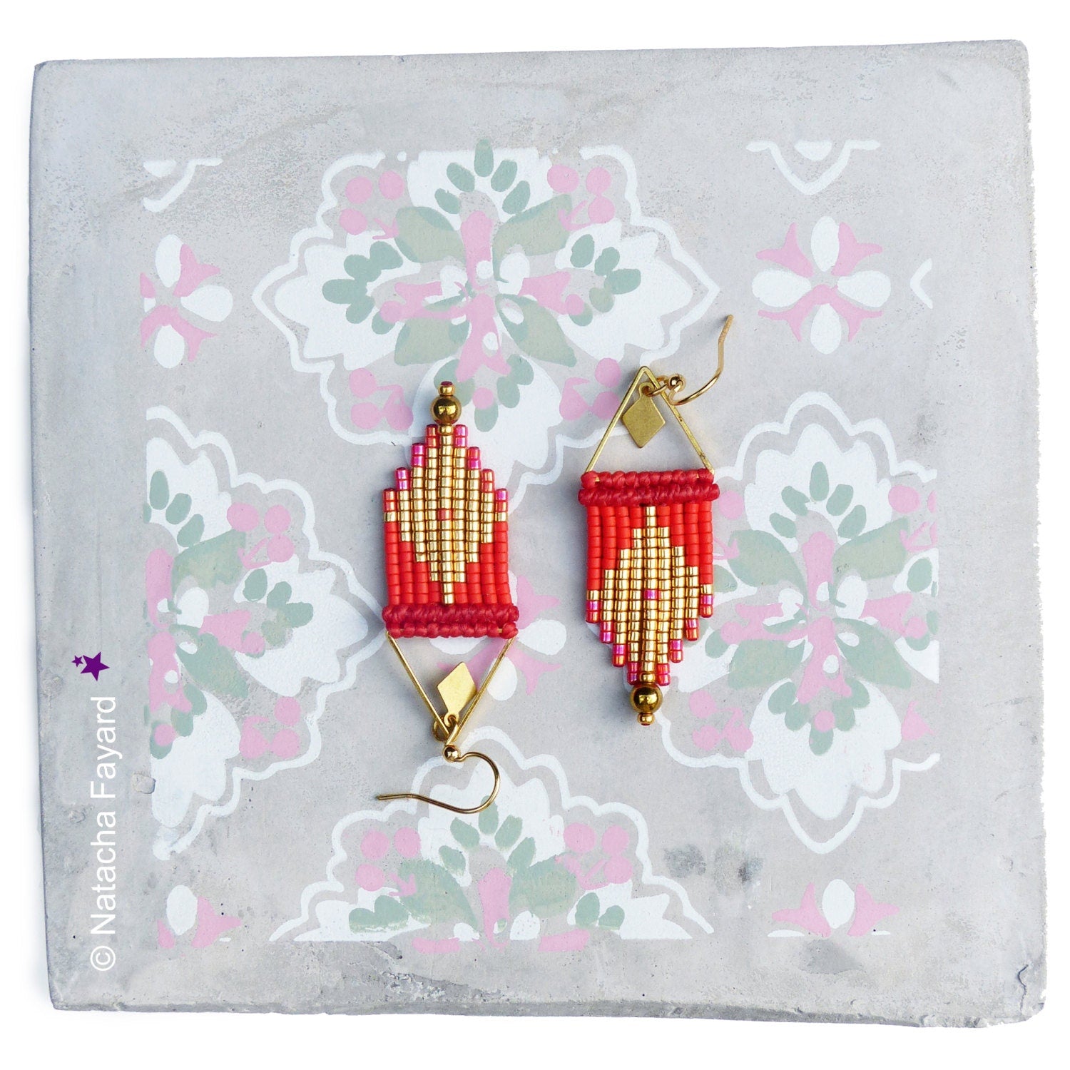 Peut inclure: Une paire de boucles d'oreilles faites à la main avec un motif géométrique. Les boucles d'oreilles présentent des perles rouges et dorées, suspendues à des montures en métal doré. Les boucles d'oreilles sont présentées sur une surface grise avec un motif floral.